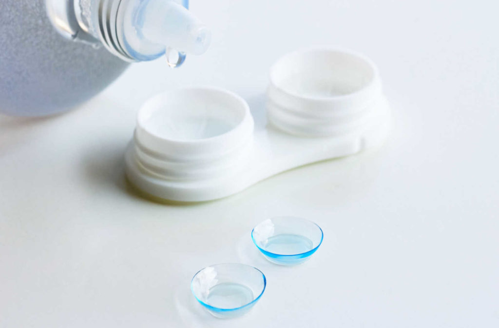 Contact lens solution drips into a contact lens case with contact lenses sitting on the table in front.