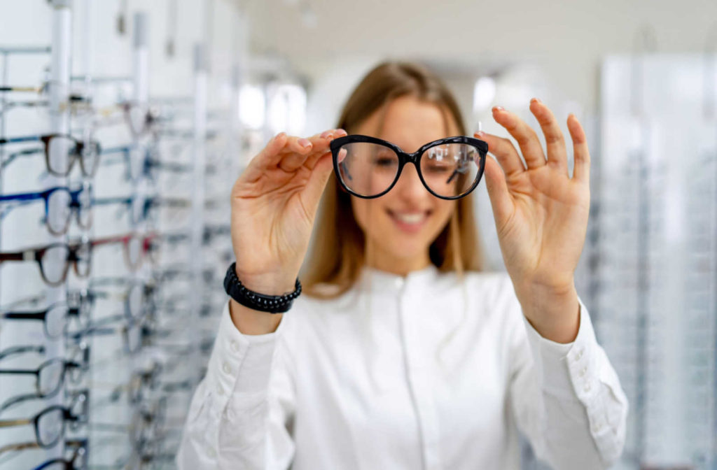 A young woman in an optical shop holds a new pair of glasses out in front of her face as she winks behind them.