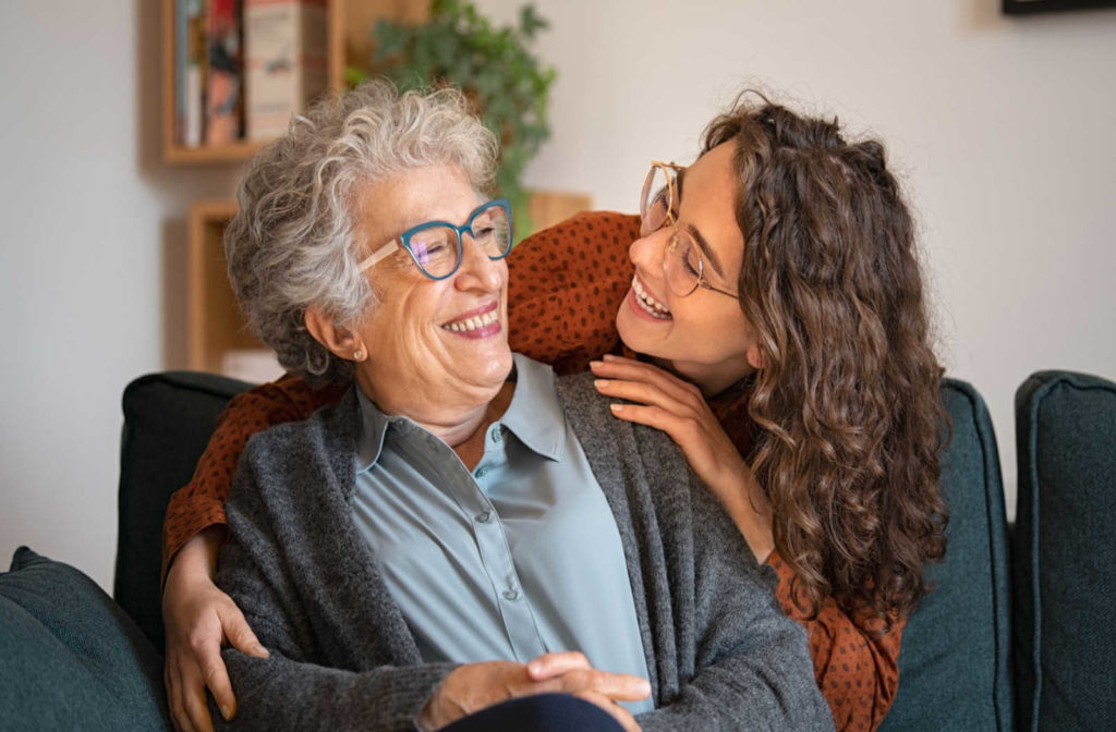 An older woman sitting on a couch wearing glasses smiles at a younger woman in glasses embracing her from behind.