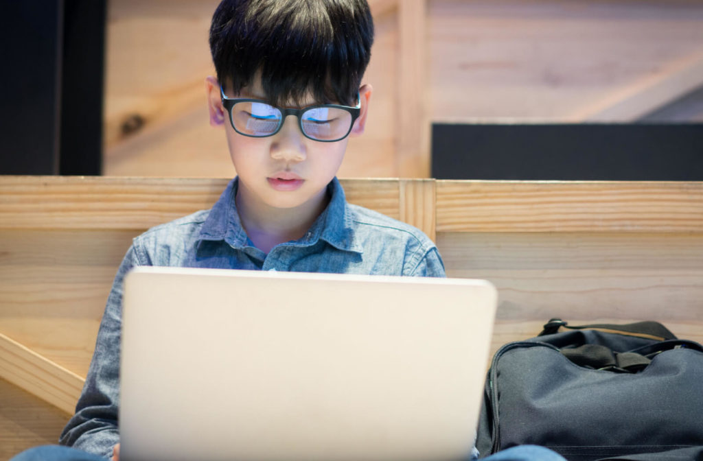 A young boy is wearing blue light glasses with studying on his computer. Blue light glasses can help to reduce or block the amount of blue light that is entering your eyes to help reduce eye strain, blurred vision, headaches, and migraines.