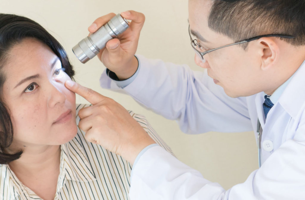 A woman facing her optometrist as he shines a small flashlight into her left eye.