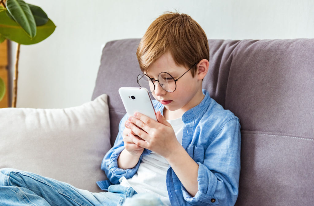 A child with eyeglasses sitting the couch and holding his smartphone very close to his face