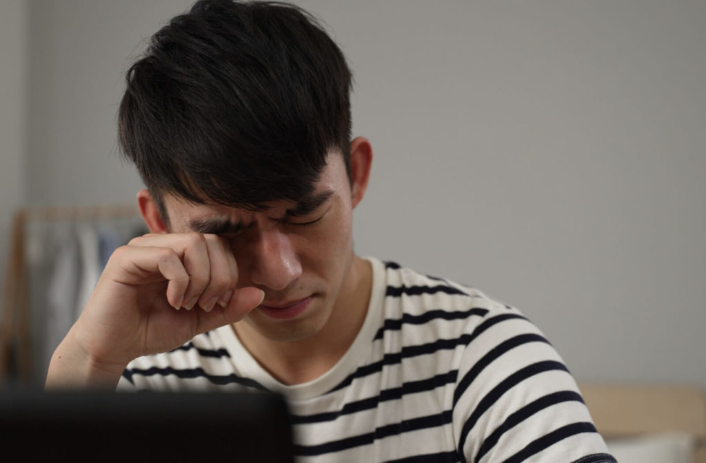 A man sitting at a desk rubbing his right eye with his fingers due to eye problems.