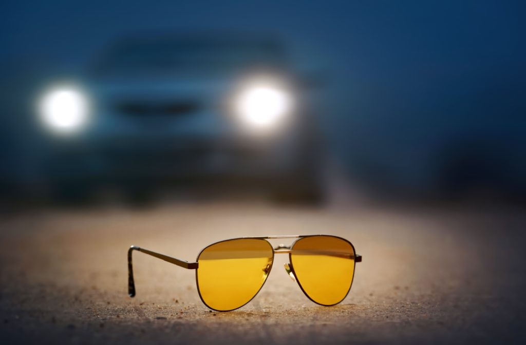 A pair of yellow tinted glasses on the ground in the foreground with a blurry car at night in the background.