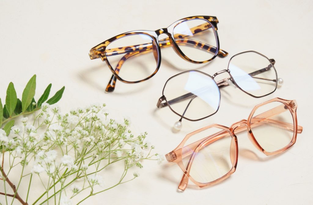 A collection of three stylish eyeglasses frames in tortoiseshell, black, and rose gold colors, arranged alongside decorative flowers on a neutral background.