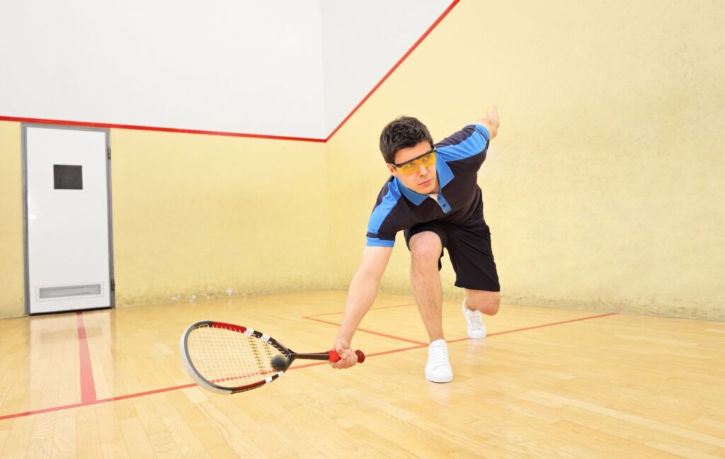 A person dives to hit a ball while playing squash. They are wearing protective glasses to protect their eyes.