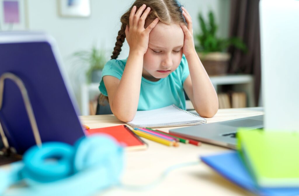 A young girl sitting at a desk, holding her head in frustration due to vision problems leading to learning difficulties.