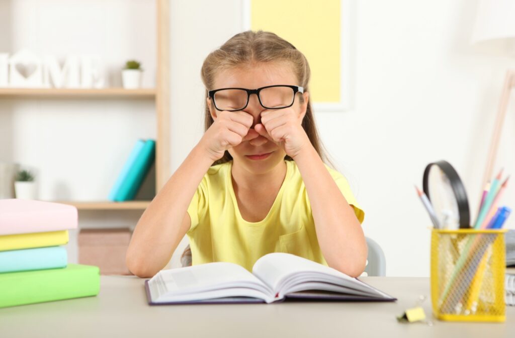 A young girl sitting at a desk, rubbing her eyes under her glasses, a sign of vision problems.