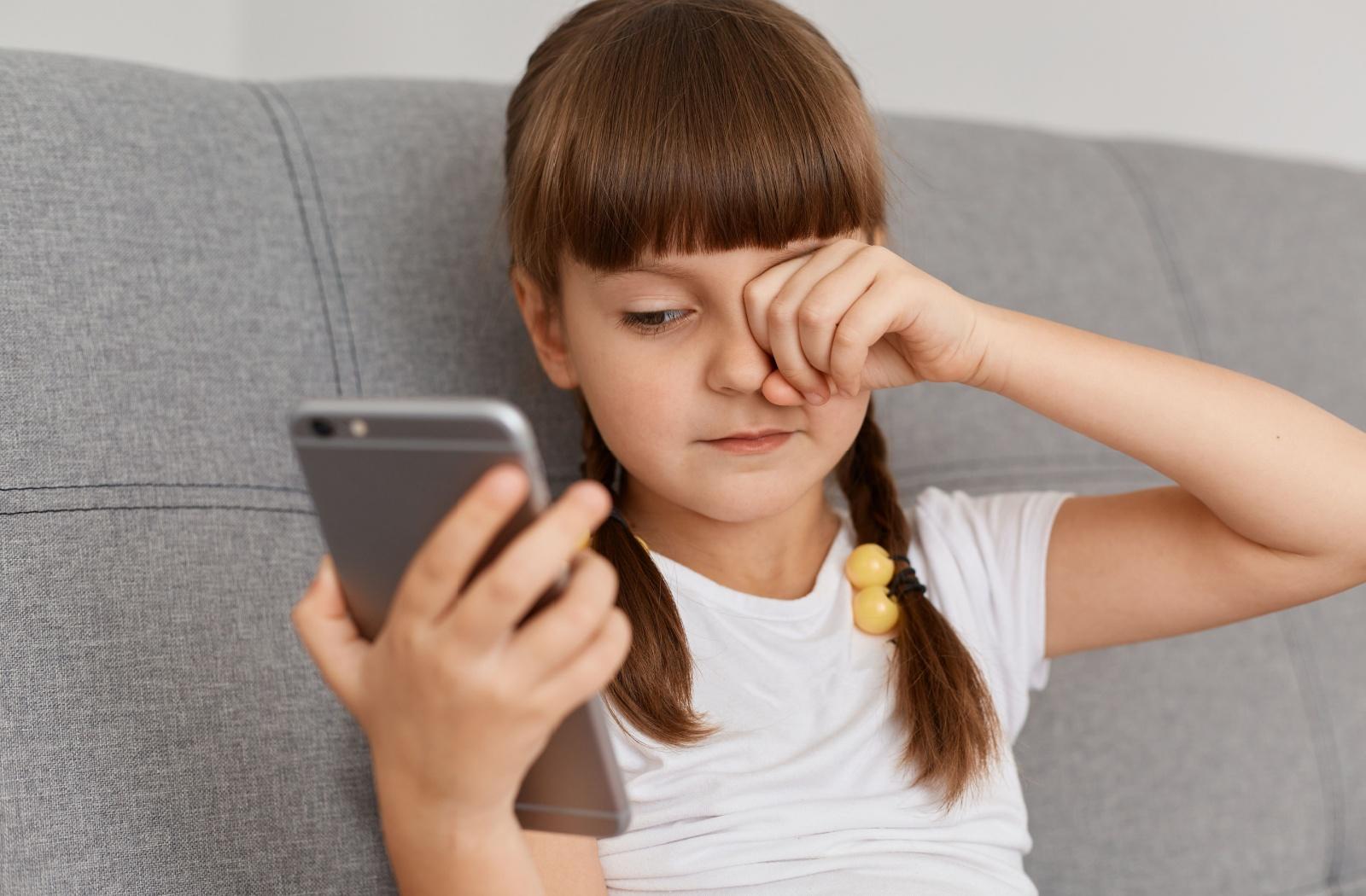 A child staring at a phone screen, rubbing their eyes.